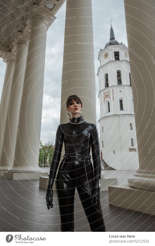 Neben der Kathedrale und dem Glockenturm von Vilnius posiert ein wunderschönes brünettes Mädchen in schwarzem Latex für einen Modeltest. Ein Modemodell trägt einen Latex-Catsuit und ist sehr hübsch. Ihr Outfit ist außergewöhnlich. Und sie auch.