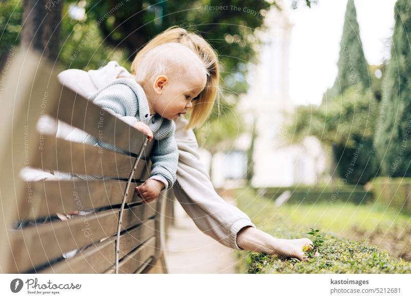 Junge Mutter mit ihrem süßen Säugling lehnt sich über die Lehne einer Holzbank in Richtung Büsche im Stadtpark und beobachtet grüne Pflanzen mit jungen Blättern und lernt über das Leben und die Natur