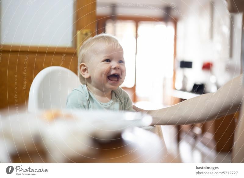 Adorable fröhliche glückliche Säugling Baby Junge Kind lächelnd beim Sitzen im Hochstuhl am Esstisch in der Küche zu Hause beeing Löffel gefüttert von seiner Mutter