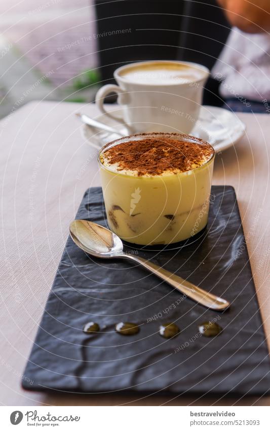 Tiramisu, ein süßes Dessert, das in Italien mit einem Löffel auf einem Tisch mit einem unscharfen Hintergrund einer Tasse heißen Cappuccino-Kaffees serviert wird.