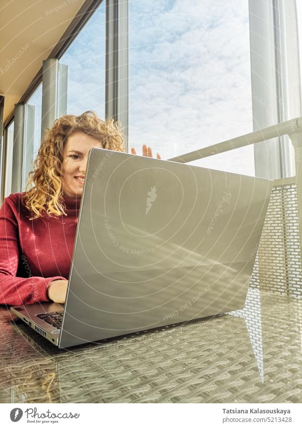 Eine Blondine mit lockigem Haar kommuniziert per Videoverbindung über einen Laptop, der vor einem Fenster steht Frau blond Behaarung Kommunizieren sitzen