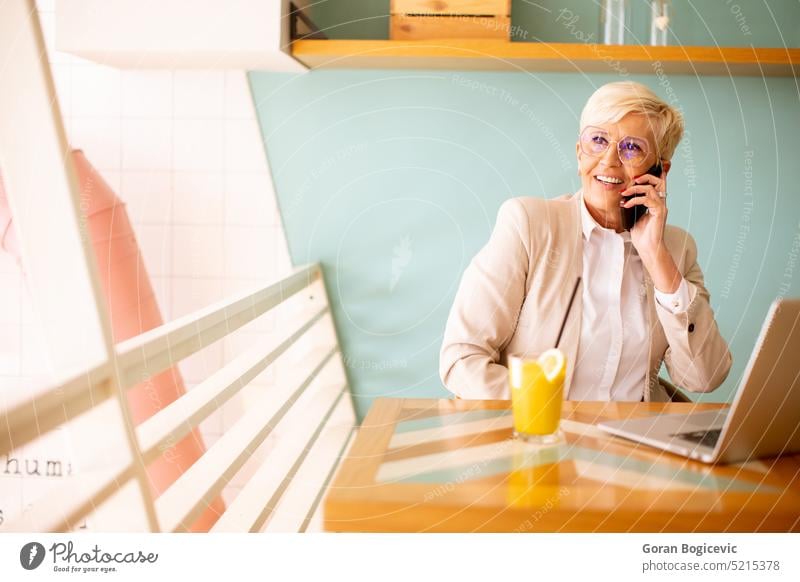 Ältere Frau, die ein Mobiltelefon benutzt, während sie am Laptop arbeitet und frischen Orangensaft im Café trinkt gealtert Kaffee Kaffeehaus Mitteilung Kunden