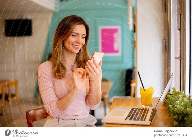 Junge Frau, die ein Mobiltelefon benutzt, während sie am Laptop arbeitet und frischen Orangensaft im Café trinkt Erwachsener attraktiv schön Schönheit Getränk