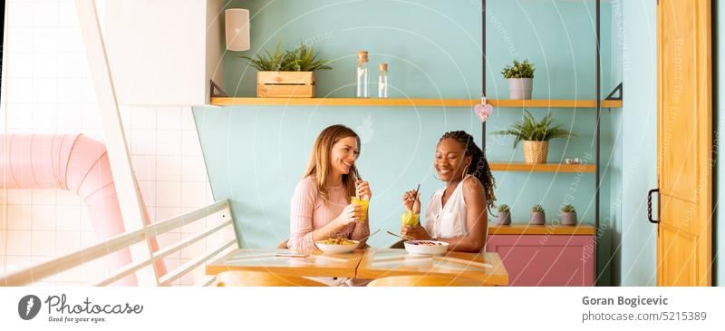 Junge schwarze und kaukasische Frau, die sich amüsiert, frische Säfte trinkt und im Café gesund frühstückt Lifestyle Gesundheit Frühstück Ethnizität Frucht