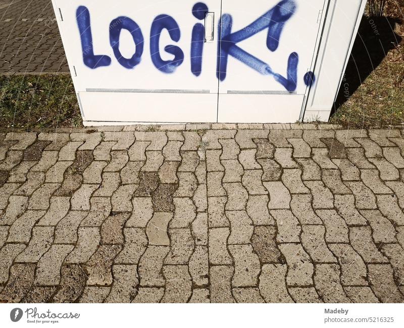 Das Wort LOGIK in blauer Schrift als Graffiti auf einem hellen Verteilerkasten am Bürgersteig mit Verbundpflaster bei Sonnenschein in Oerlinghausen bei Bielefeld am Hermannsweg im Teutoburger Wald in Ostwestfalen-Lippe