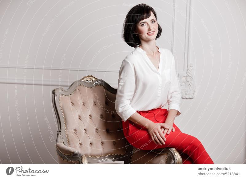 Studio Porträt der attraktiven jungen Frau mit dunklen Haaren in weißem Hemd und roten Hosen sitzen auf klassischen Stuhl auf schlichten weißen Hintergrund.bossy weiblich, Vorstellungsgespräch Konzept, selbstbewusste unabhängige Dame