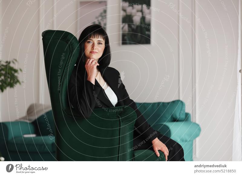 Schöne Geschäftsfrau in schwarzem Anzug und weißer Bluse sitzt auf dem grünen Sofa im Büro, charmante brünette Frau mit kurzen Haaren schaut in die Kamera, Mädchen arbeitet drinnen