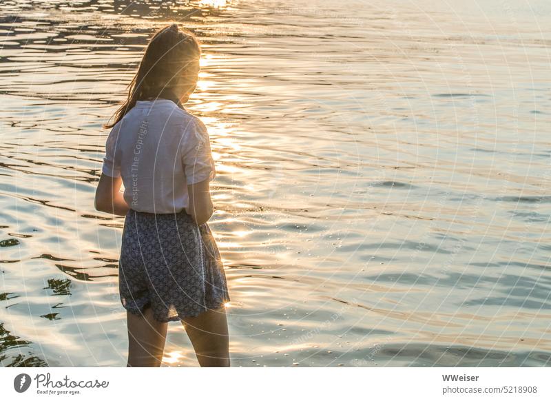Eine junge Frau steht mit den Füßen im Wasser und beobachtet das Glitzern der Wellen Mädchen Sonne Licht Sonnenuntergang warm glitzern strahlen weich See