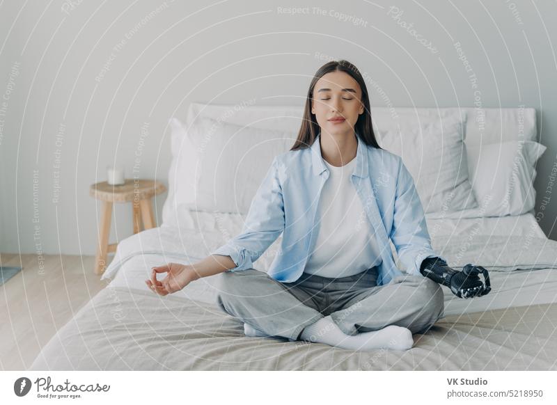 Behindertes Mädchen mit bionischer Armprothese übt Yoga und meditiert im Lotussitz auf dem Bett zu Hause deaktiviert Prothesen meditieren atmen Lotos Wellness