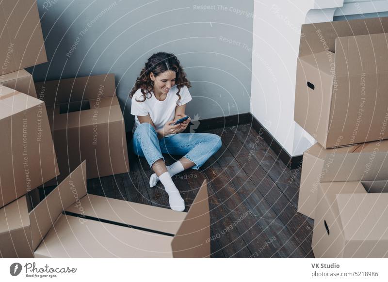 Modernes Mädchen sitzt mit Kisten und nutzt Smartphone-App, um Umzugsunternehmen und Spediteur für den Umzug auszuwählen Frau Faltschachtel bewegend Schachtel