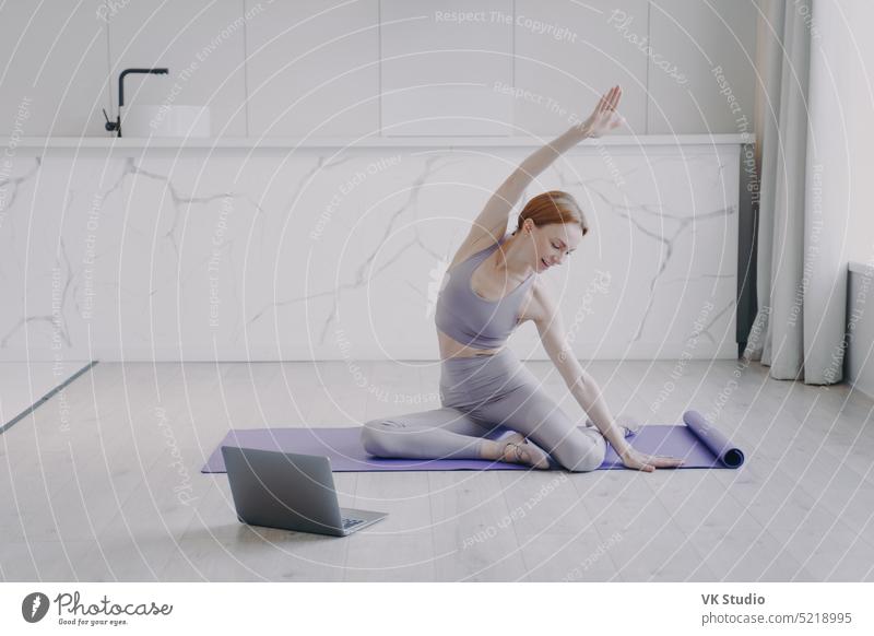 Junge Frau dehnt sich und übt Yoga auf dem Boden. Sport und gesunder Lebensstil auf Quarantäne. Person Laptop Computer Pilates Tutorial trainiert. Lernen