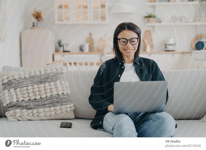Lächelnde Frau, die am Laptop arbeitet, in einem Online-Shop einkauft und auf einer gemütlichen Couch zu Hause sitzt. E-Commerce Arbeit Liege heimwärts
