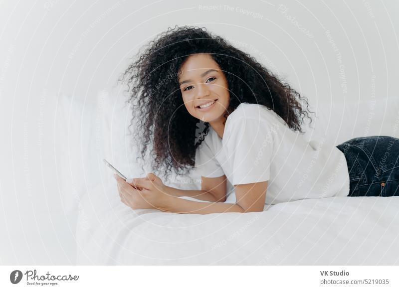 Hübsche fröhliche Frau mit Afro-Haarschnitt, genießt Chatten und Networking, liegt auf einem bequemen Bett, schaut Fotos, hat entspannten Gesichtsausdruck, isoliert auf weißem Hintergrund. Online-Kommunikation