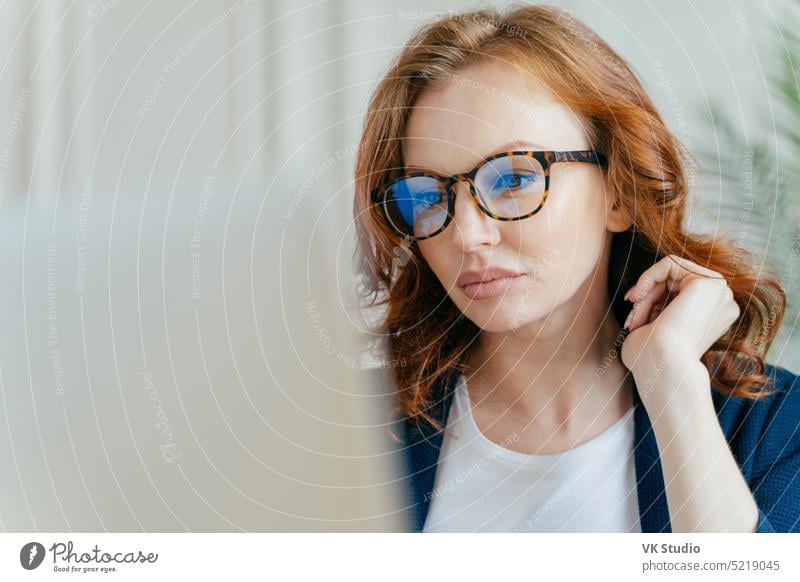 Nahaufnahme einer fokussierten weiblichen Arbeitskraft, die ein erfolgreiches Projekt abschließt, eine Fernarbeit macht, sich auf den Monitor eines Laptops konzentriert, eine optische Brille trägt, um die Sicht zu korrigieren, ingwerfarbene lockige Haare hat