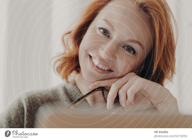 Close up shot von glücklichen schönen rothaarige Frau neigt den Kopf, lächelt positiv, hat weiße perfekte Zähne, Make-up, sitzt vor dem geöffneten Laptop-Computer, sucht notwendige Informationen, nutzt kostenlose wifi