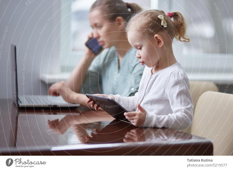 Ein Mädchen spielt mit einem Tablet. Gestresste Mutter bei der Arbeit, die einen Anruf entgegennimmt und sich um ihr Baby im Büro kümmert Kind Apparatur