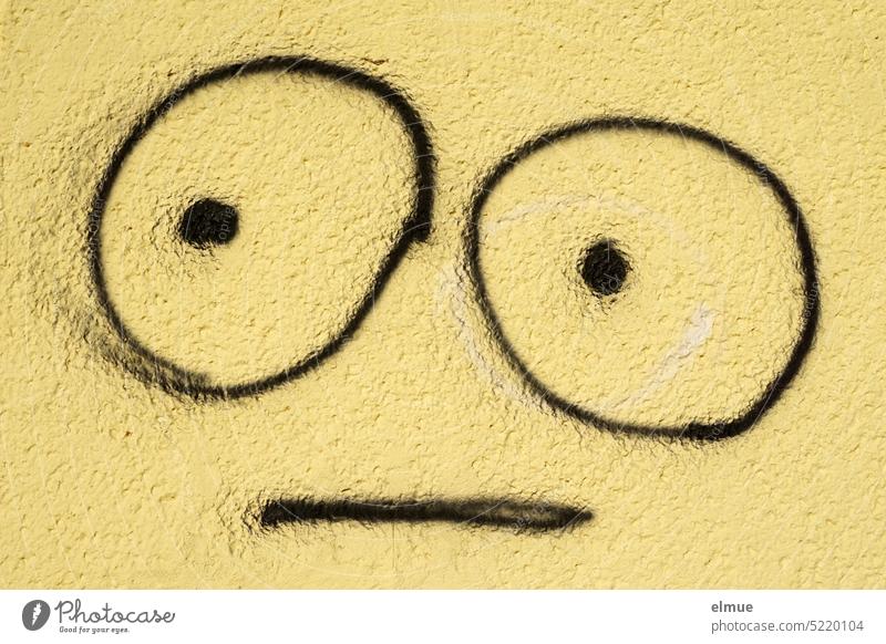 an eine gelbe Wand mit schwarz gezeichnetes Emoji mit großen Augen und neutralem Ausdruck Fratze Grimasse Smiley Smiley-Zeichnung neutraler Blick zeichnen