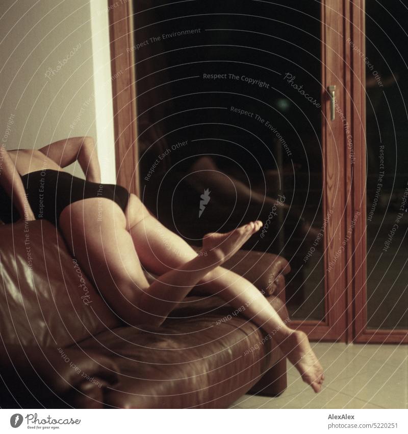 schlanke, sportliche Frau in schwarzem Body kniet mit einem Bein auf einer Couch über die Lehne und hält die Beine in die Luft vor einem Balkonfenster - analoges, rechteckiges Foto