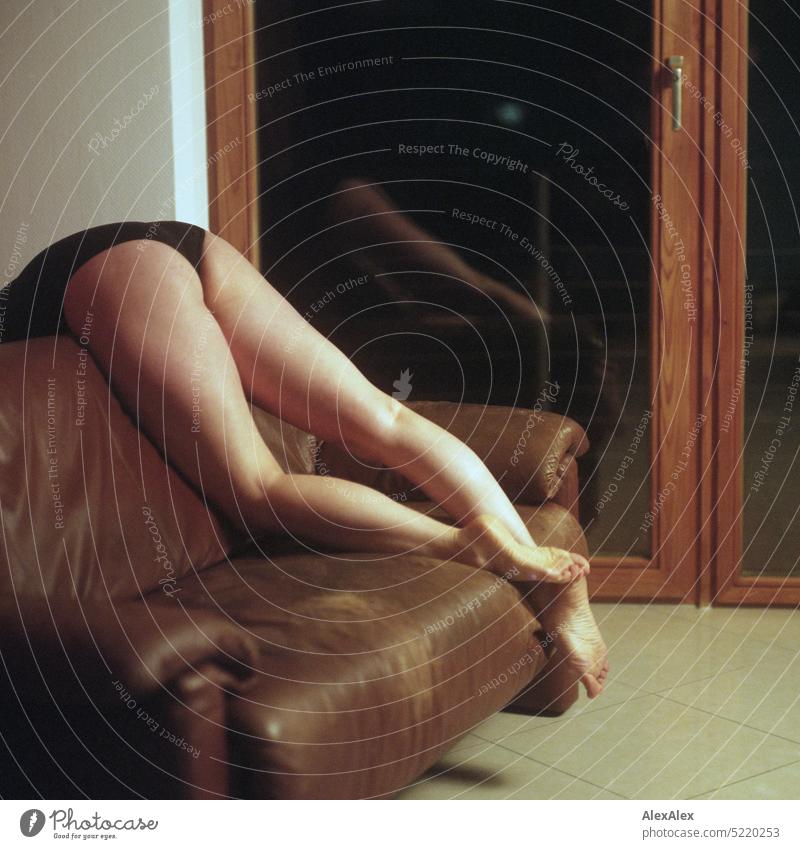 schlanke, sportliche Frau in schwarzem Body liegt auf einer Couch über der Lehne und hält die Beine in die Luft vor einem Balkonfenster - analoges, rechteckiges Foto