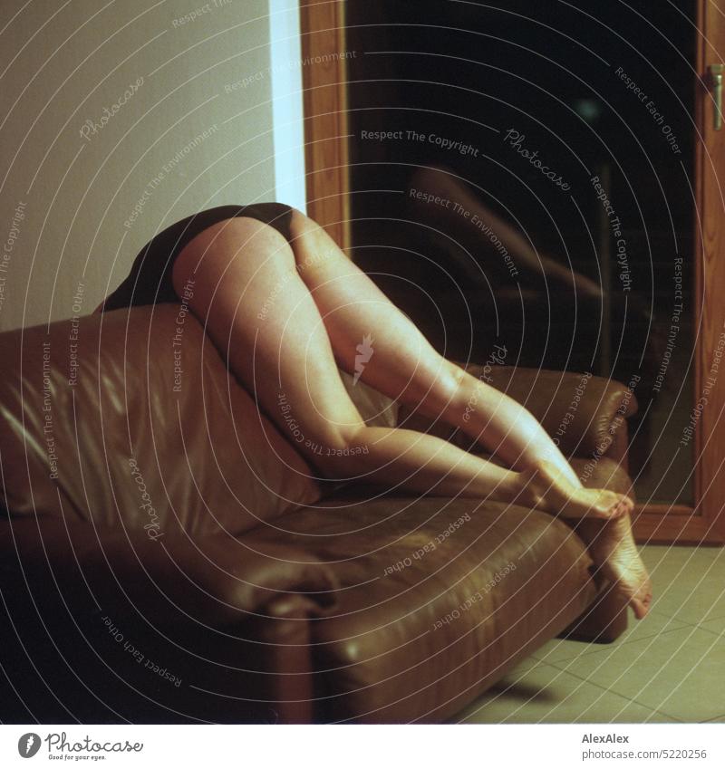 schlanke, sportliche Frau in schwarzem Body liegt auf einer Couch über der Lehne und hält die Beine in die Luft vor einem Balkonfenster - analoges, rechteckiges Foto