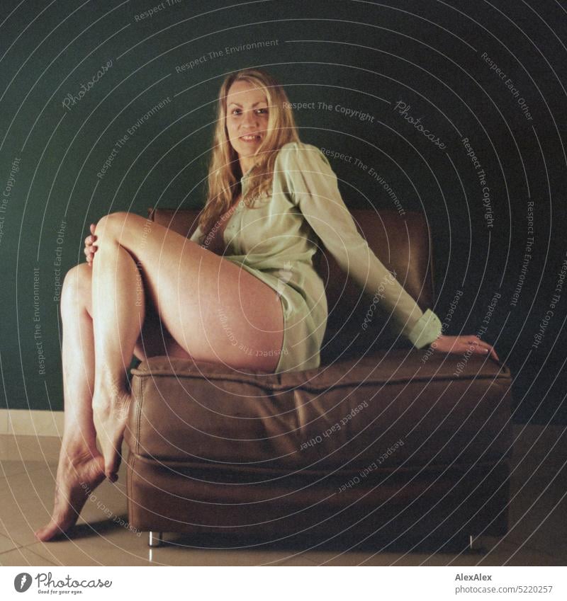 schöne, blonde Frau mit nackten Beinen und durchscheinender Bluse sitzt quer auf einem braunen Sessel vor einer türkisen Wand Haut Barfuß kraftvoll sexy Abend