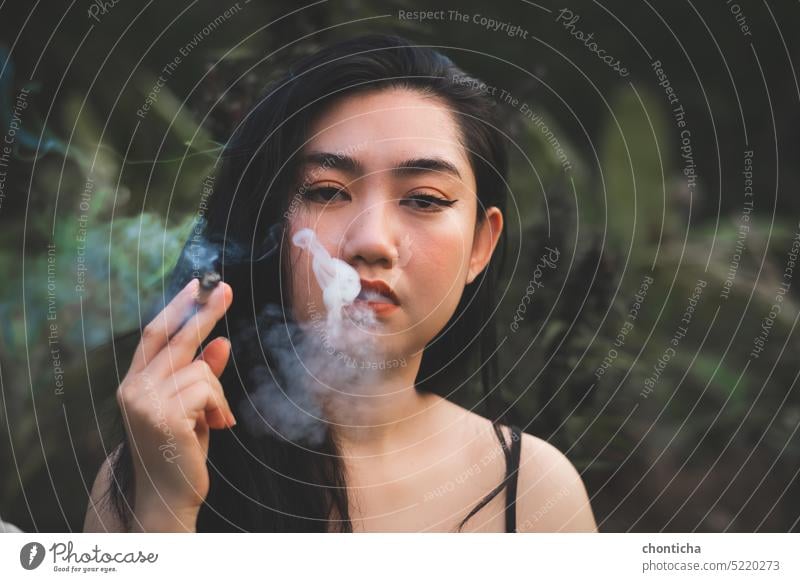 Yong Asia Frau raucht an Unschärfe Baum Pflanze Hintergrund Süchtige Erwachsener Kunst attraktiv schön Schönheit schwarz Cannabis Kaukasier heiter Zigarette