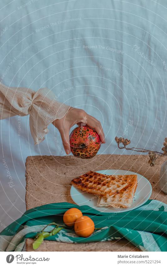Vertikale verträumte Aufnahme einer Hand, die einen Granatapfel über einem Tisch mit Mandarinen und Waffeln zum Frühstück hält, Konzept des Frühlings, des häuslichen Lebens, der Wellness und des langsamen Lebensstils