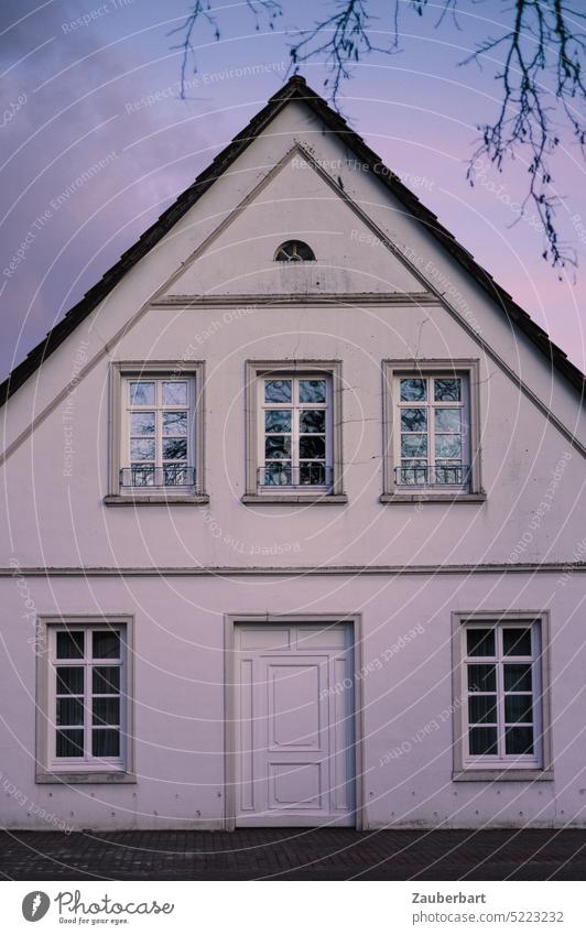 Weiße Giebelseite eines alten Hauses mit Satteldach und Sprossenfenstern im violett gefärbten Abendlicht, symbolisch für Bestandshaus, renovieren und dämmen