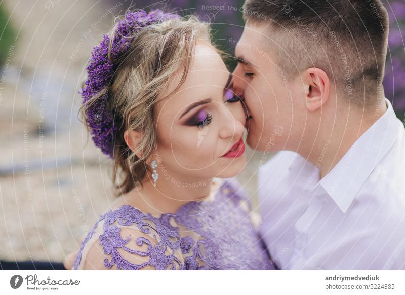 Junges verliebtes Paar küsst sich in einem Lavendelfeld an einem sonnigen Sommertag. Mädchen in einem luxuriösen lila Kleid und mit Frisur Liebe Menschen