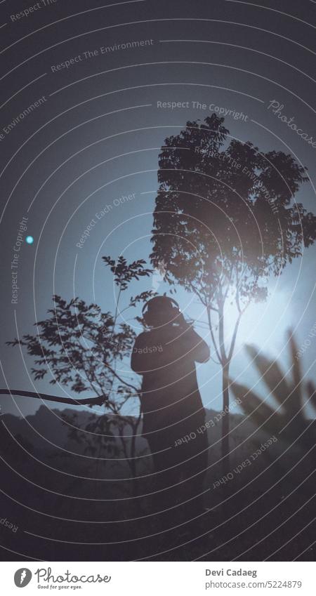 Eine Silhouette einer Frau mit Kopfhörern, die ein Foto unter der Sonne macht Himmel Baum blau Ast Baumstamm Natur Wolken Sommer grün Fotokamera fotografierend