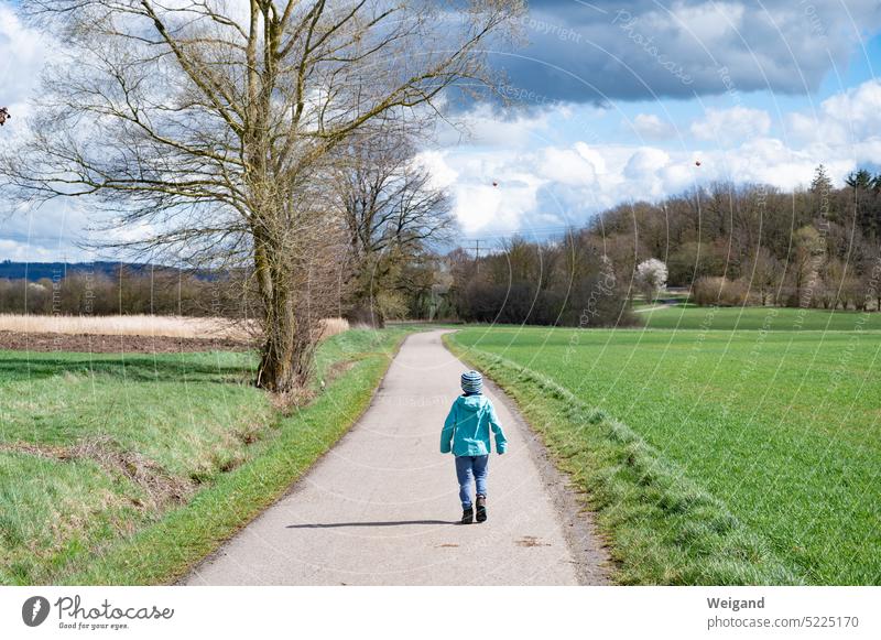 Kleines Kind spaziert auf einem Feldweg durch grüne Wiesen bei Frühjahrswetter und blauem, bewölktem Himmel mit Wald in der Ferne Natur Spaziergang Grün saftig