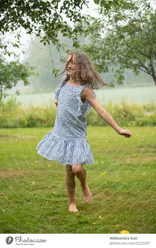 Glücklich niedlichen kleinen Mädchen im Sommerkleid tanzen und laufen auf grünem Rasen unter dem regen. Barfuß Kind genießt den Sommer im Freien. schön