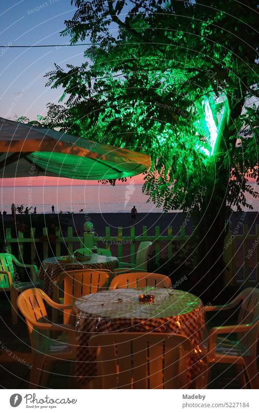 Terrasse eines Café mit Plastikstühlen und Tischdecke aus Wachstuch mit grüner Beleuchtung nach Sonnenuntergang am Strand von Inkumu am Schwarzen Meer bei Bartin an der Schwarzmeerküste in der Türkei