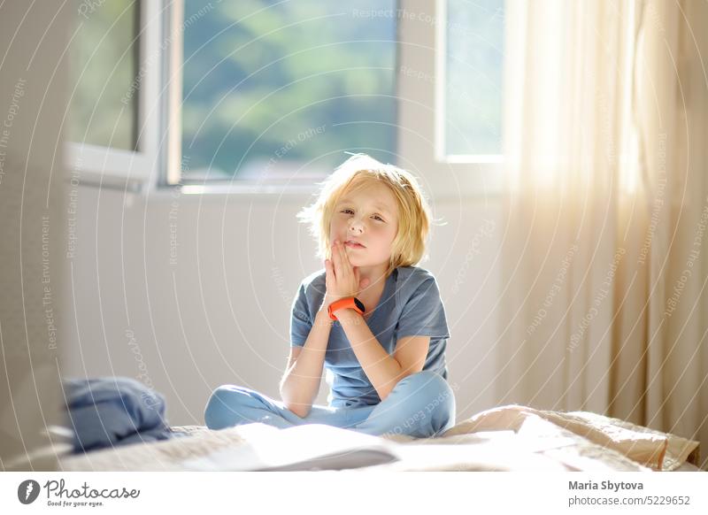 Porträt eines niedlichen Schuljungen in einem blauen T-Shirt im Schlafzimmer an einem sonnigen Tag. Preteen Junge sitzt auf dem Bett und träumt. Kind
