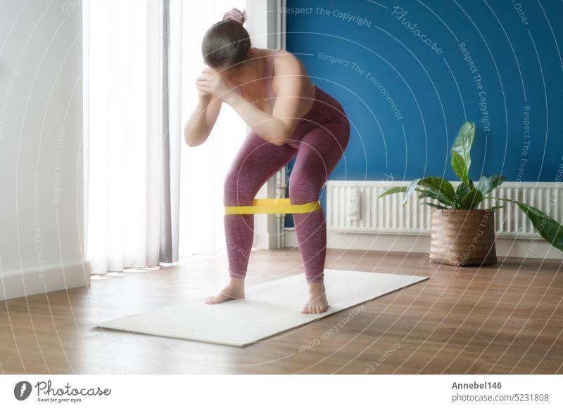 Frau während ihres Fitnesstrainings zu Hause mit Gummiwiderstandsband am Bein bei Kniebeugen Sport heimwärts Training Lifestyle Sportbekleidung Gesundheit Übung