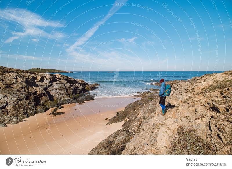 Rucksacktourist wandert entlang eines Felsens und über einen Sandstrand auf dem markierten Fisherman Trail an der Atlantikküste bei Porto Covo, Portugal. Rota Vicentina