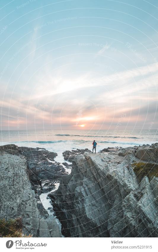 Ein furchtloser Mann mit einem Rucksack beobachtet den Sonnenuntergang über dem Atlantik in Porto Covo, Portugal. Ein Wanderer steht am Rande einer Klippe und schaut in die Zukunft. Selbsterkenntnis