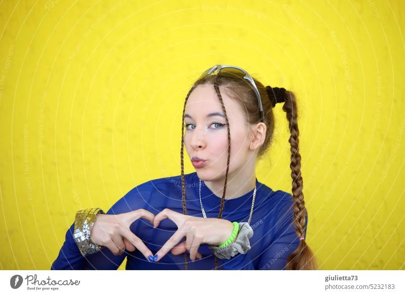 Cooles Teenager Girl, gestylt mit Zöpfen, süßem Kussmund und mit den Fingern ein Herz zeigend vor gelber Wand 13-18 Jahre 18-20 Jahre 17 Jugendliche Girly