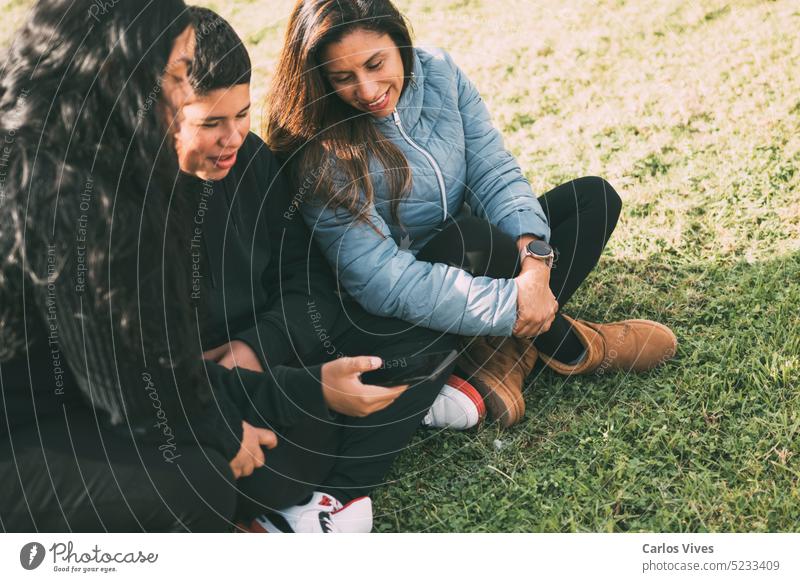 Eine hispanische Familie verbringt an einem schönen, sonnigen Tag Zeit miteinander in einem örtlichen Park. Ein Teenager sitzt im Gras, hält sein Smartphone in der Hand und schaut weg. Seine Mutter und seine Schwester sitzen neben ihm, schauen in die Kamera und lächeln.