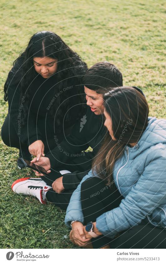 drei Personen hispanisch-lateinamerikanischer Ethnizität, die mit intelligenten Geräten auf dem Boden im Park sitzen Aktivität Atmosphäre authentisch Browsen