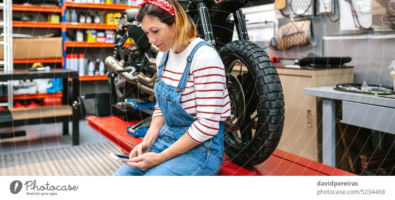 Mechanikerin mit Blick auf ein Telefon, das auf einer Plattform mit einem Motorrad in einer Fabrik steht Frau benutzend Funktelefon Lächeln Fröhlichkeit Porträt