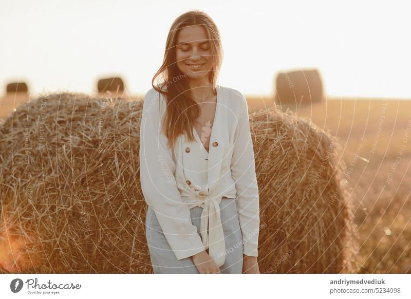 Porträt eines lächelnden schönen Mädchens mit langen Haaren in einem Jeansrock. Frau, die einen Spaziergang in einem Weizenfeld mit Heuballen an einem sonnigen Sommertag genießt.