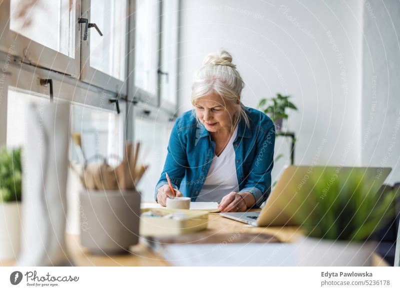 Lächelnde reife Geschäftsfrau, die in ein Notizbuch schreibt, während sie am Tisch im Büro sitzt echte Menschen Senior im Innenbereich Loft Fenster heimwärts