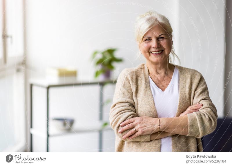 Porträt einer lächelnden älteren Frau, die mit verschränkten Armen steht echte Menschen Senior im Innenbereich Loft Fenster heimwärts reifer Erwachsener