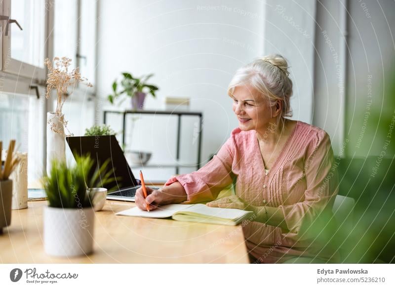 Lächelnde reife Geschäftsfrau, die in ein Notizbuch schreibt, während sie am Tisch im Büro sitzt echte Menschen Senior im Innenbereich Loft Fenster heimwärts