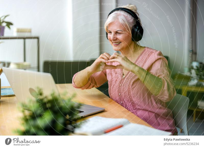 Ältere Frau formt ein Herz mit ihren Händen während eines Video-Chats echte Menschen Senior im Innenbereich Loft Fenster heimwärts reifer Erwachsener