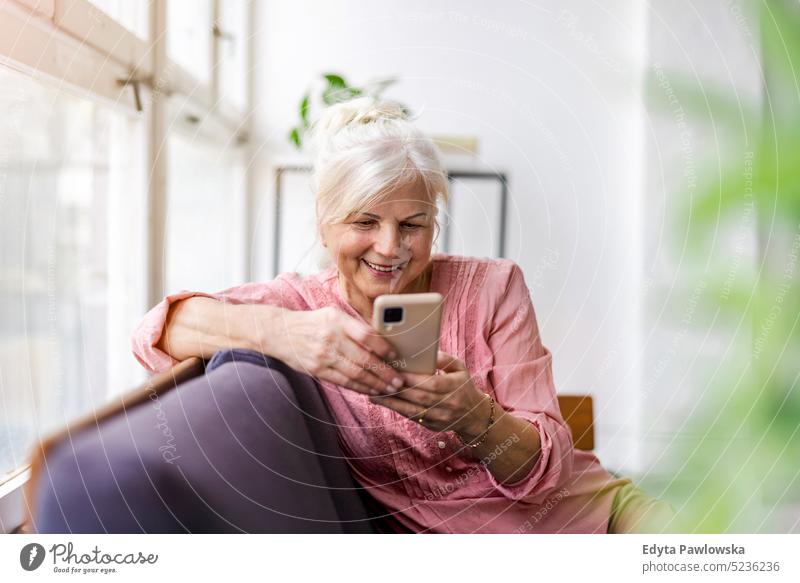 Lächelnde ältere Frau mit Smartphone echte Menschen Senior im Innenbereich Loft Fenster heimwärts reifer Erwachsener eine Person attraktiv gelungen