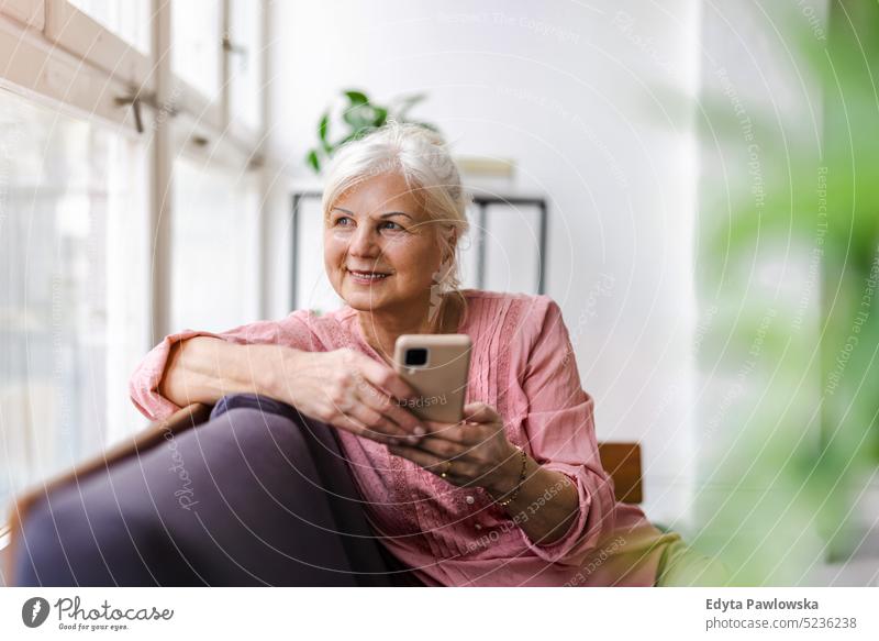 Lächelnde ältere Frau mit Smartphone echte Menschen Senior im Innenbereich Loft Fenster heimwärts reifer Erwachsener eine Person attraktiv gelungen