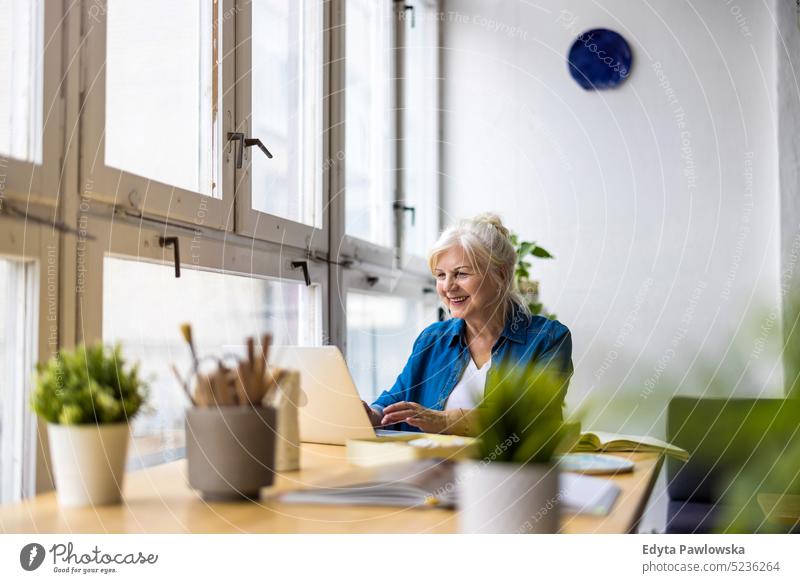 Porträt einer lächelnden reifen Geschäftsfrau mit Laptop am Schreibtisch im Büro echte Menschen Senior im Innenbereich Loft Fenster heimwärts reifer Erwachsener