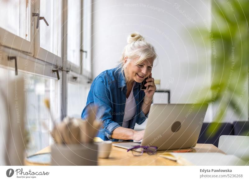 Lächelnde Geschäftsfrau, die mit dem Handy telefoniert und einen Laptop im Büro benutzt echte Menschen Senior im Innenbereich Loft Fenster heimwärts